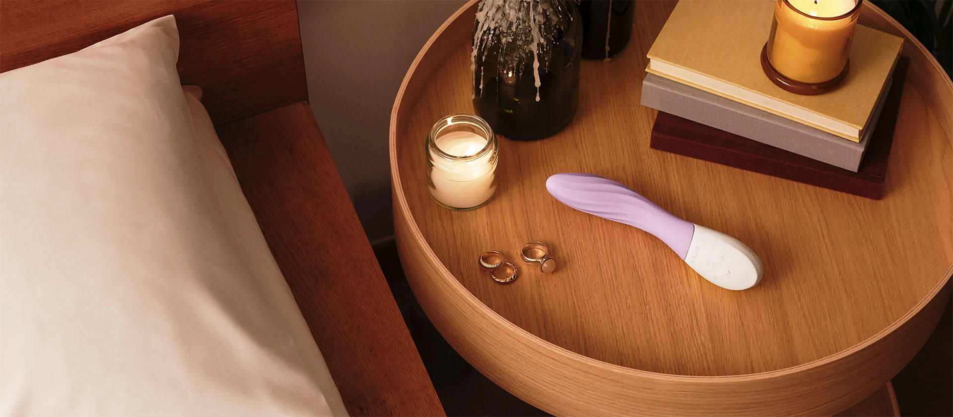 Lavender LELO MONA Twist G-spot vibrator displayed in a warm, lit bedroom nightstand setting.